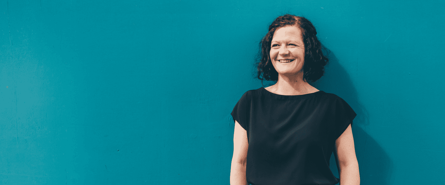 A woman with short dark hair stands in front of a teal wall, smiling.