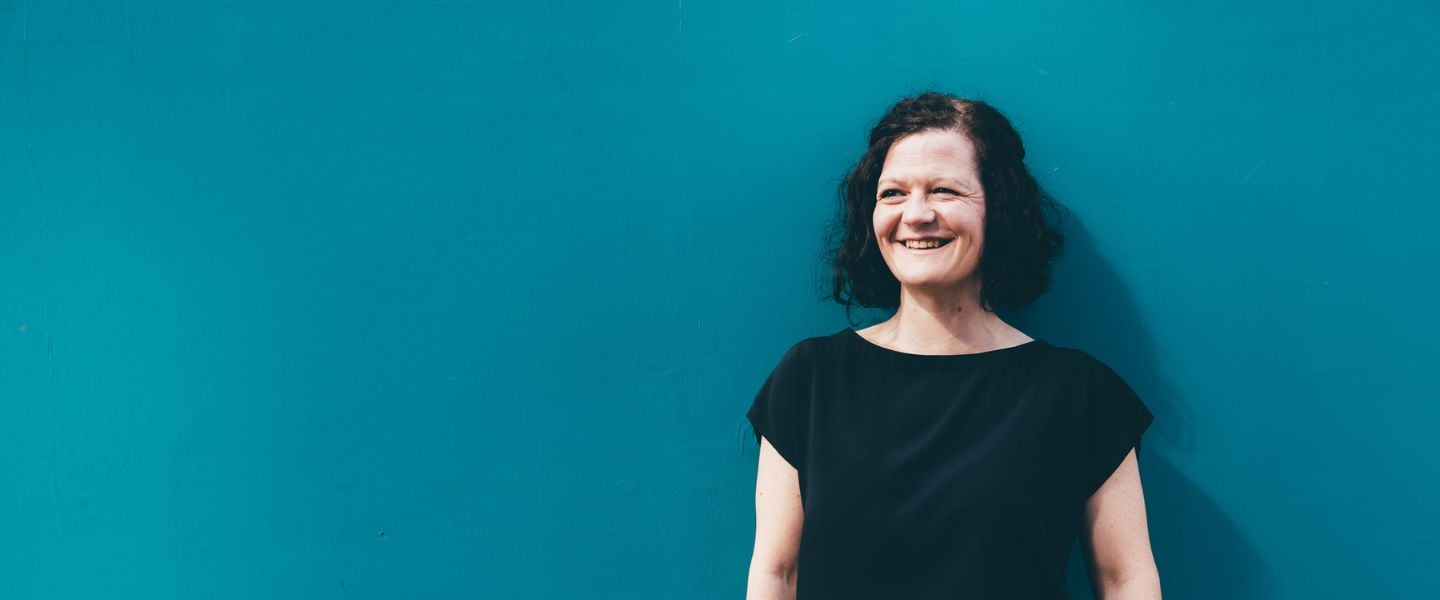 A woman with shoulder length brown hair pictured wearing a black top, standing in front of a bright teal wall.