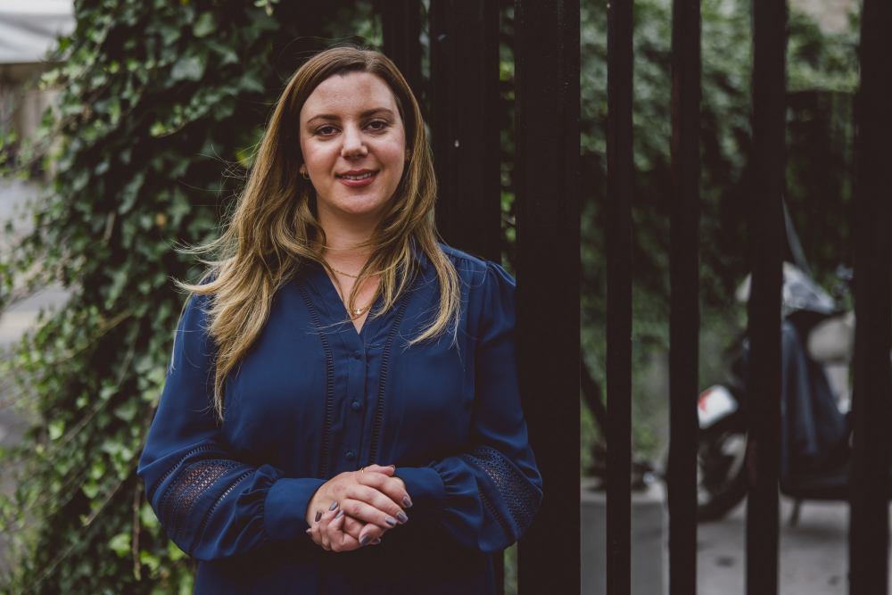 A woman in a blue blouse stands in front of a fence, with ivy in the background. She is smiling softly and appears relaxed.