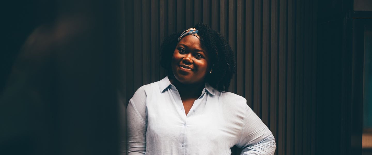 A woman wearing a pale blue shirt, smiling at the camera with a hand on her hip. She is photographed outside, against a black brick wall and black glass wall.