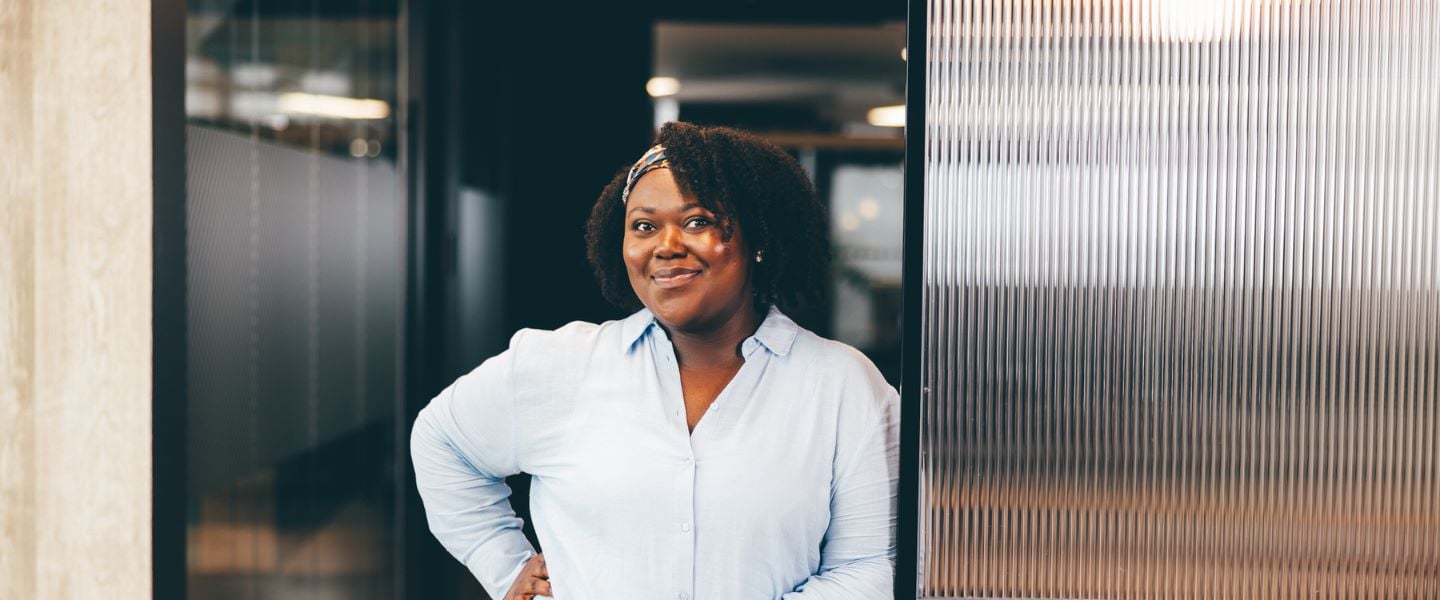 A woman is pictured in an office space, wearing a pale blue shirt and white trousers. She is smiling at the camera with a hand on her hip.