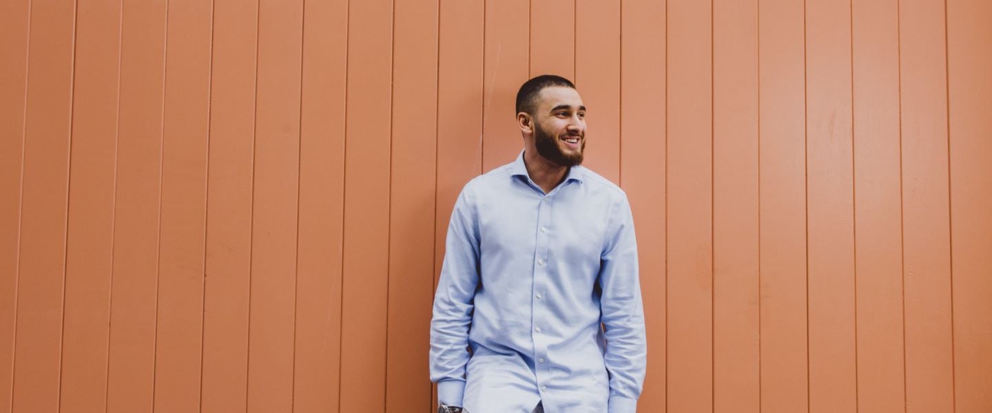 A young man is stood leaning against a peach coloured wooden high wooden door. He is wearing a pale blue shirt, and smiling whilst looking off camera to the left.