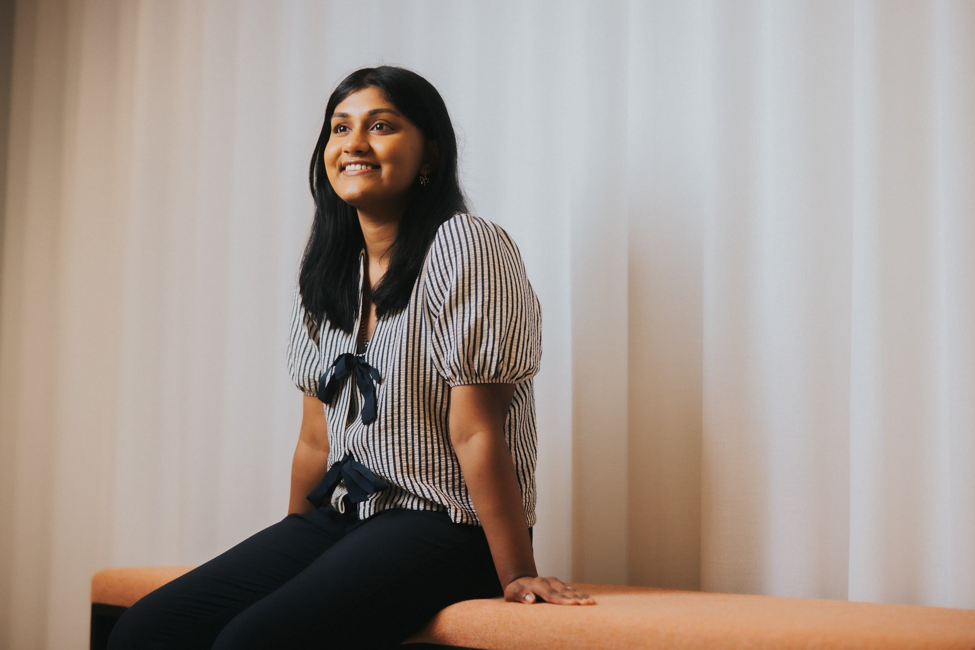 A young Asian woman is sitting on a comfy bench seat inside an office building. She has long black hair and is wearing a black and white striped blouse and black trousers. She is looking away from the camera and smiling.