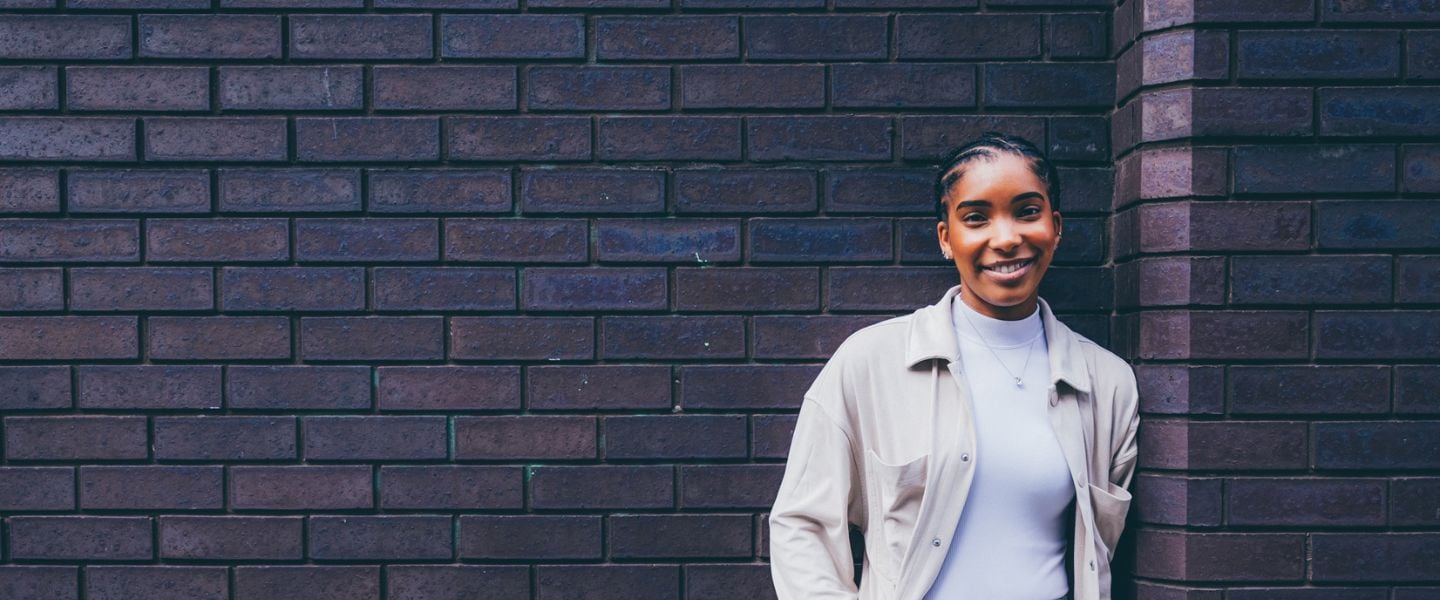 A young woman is pictured leaning against a dark brick wall. She is looking directly at the camera and smiling, wearing grey tailored trousers and a pale cream top and shirt.