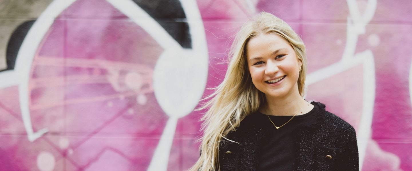 A young woman with long blond hair is photographed wearing a black top, stood against a fence in front of bright pink graffiti.