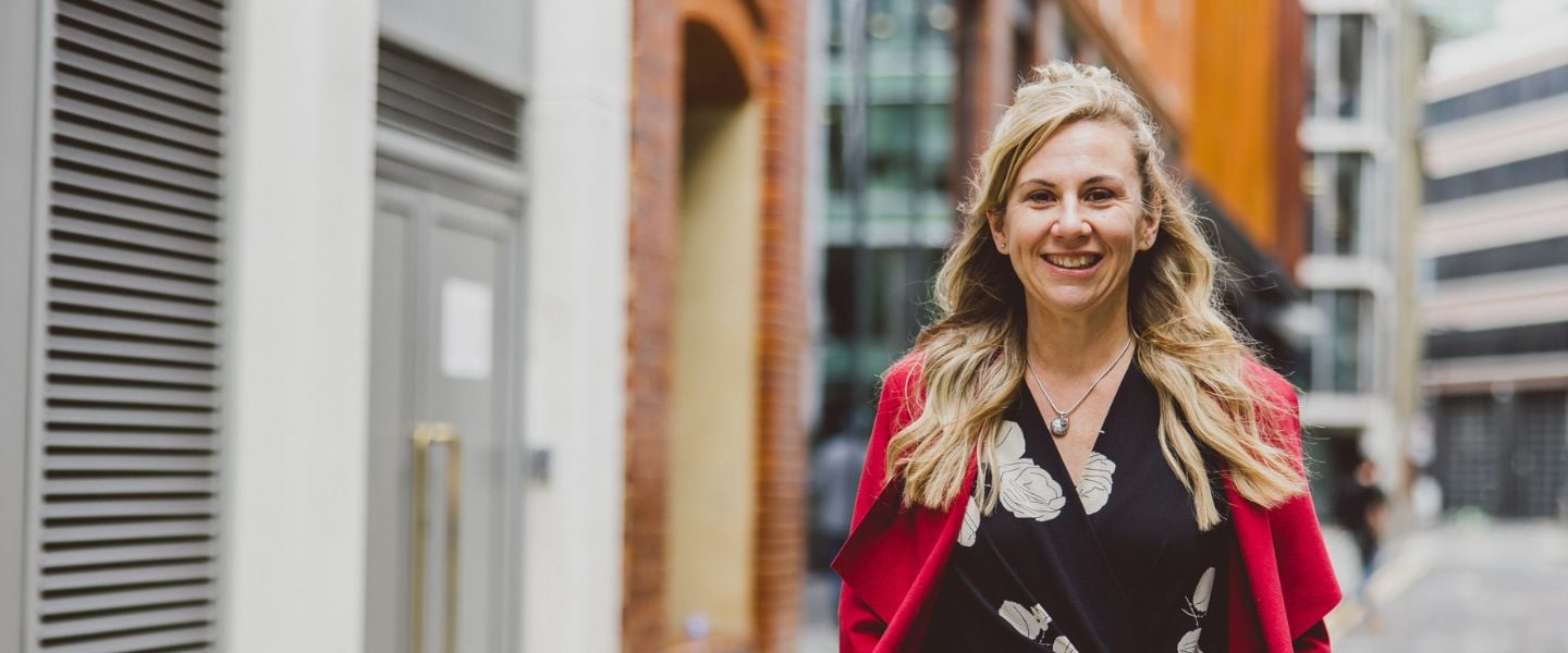A woman with long blond hair is pictured walking towards the camera, smiling. She is wearing a black and white top with a bright red jacket.