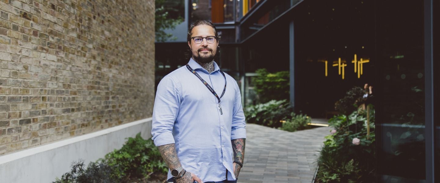A man is smiling and looking directly at the camera, with his hands tucked into his jean pockets. He is wearing a pale blue shirt, and a lanyard signalling neurodiversity. He has glasses and you can see tattoos at the top of his shirt and on his arms, and two piercings on his chin. He is standing in front of a pale brick wall.