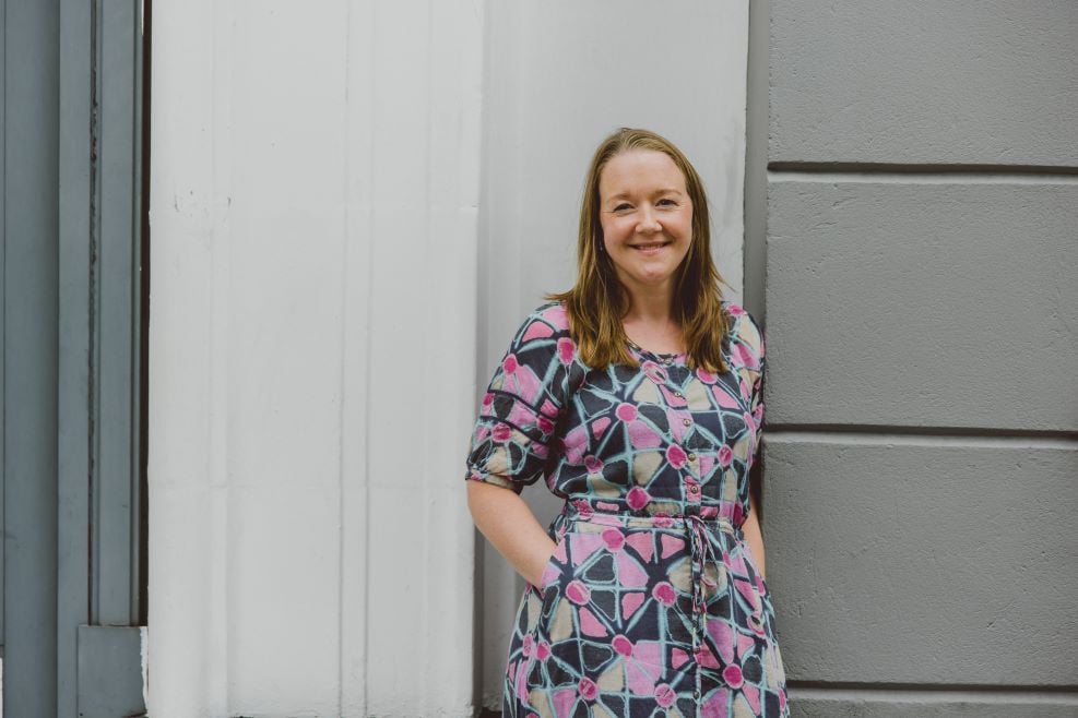 A lady is pictured standing in front of a grey and white brick wall. She is smiling at the camera and wearing a grey dress with brightly coloured patterns.