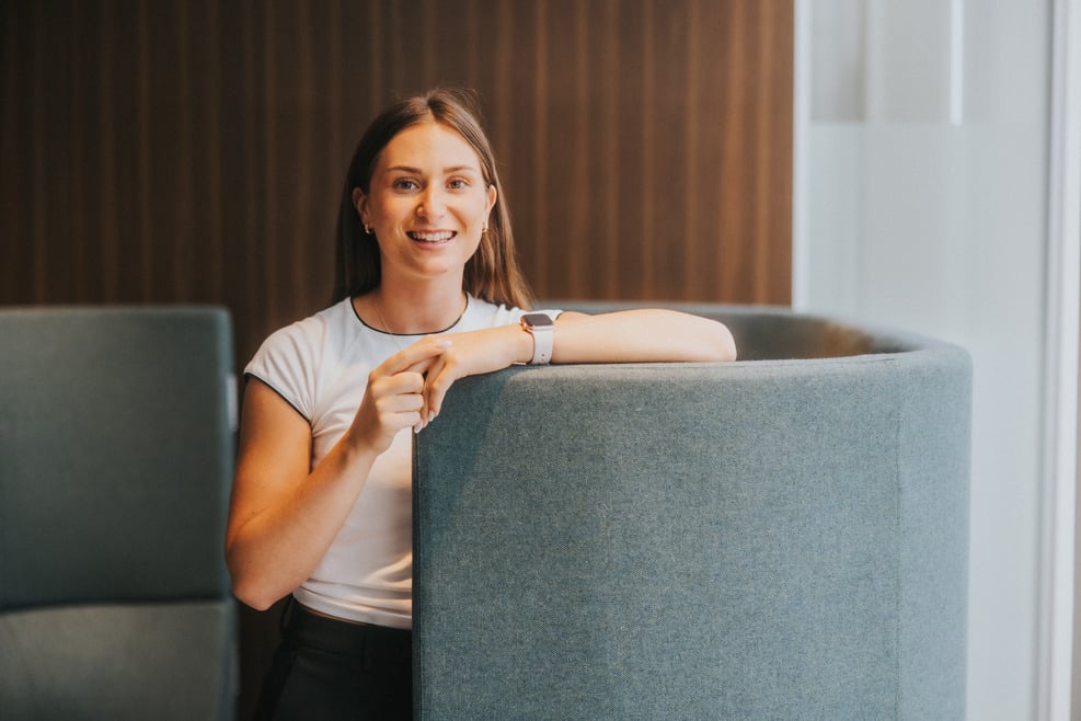 A woman with long brown hair leans her arm on top of a green chair, wearing a white t-shirt and black trousers. She's smiling at the camera.