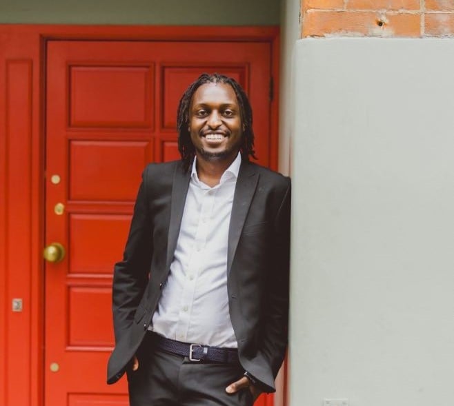 A man with his hair in braids is wearing an open suit jacket and white shirt, smiling and stood in front of a red door.