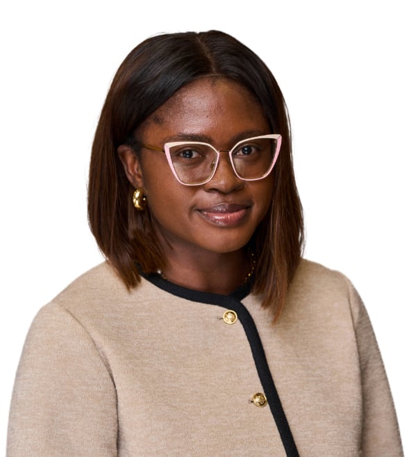 A young Black woman with short brown hair is posing for a headshot with only her face and shoulders visible. She is smiling and wearing a pair of large glasses and beige jacket.
