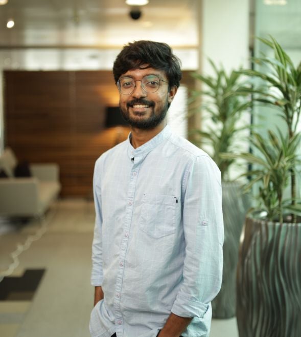 A man with glasses and a beard, wearing a light blue shirt, stands indoors in a modern office space, smiling at the camera.