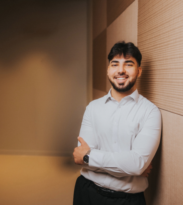 A young man with short dark brown hair is smiling to the camera. He is standing inside and is learning against a beige coloured wall. He is wearing a formal white shirt and black trousers.
