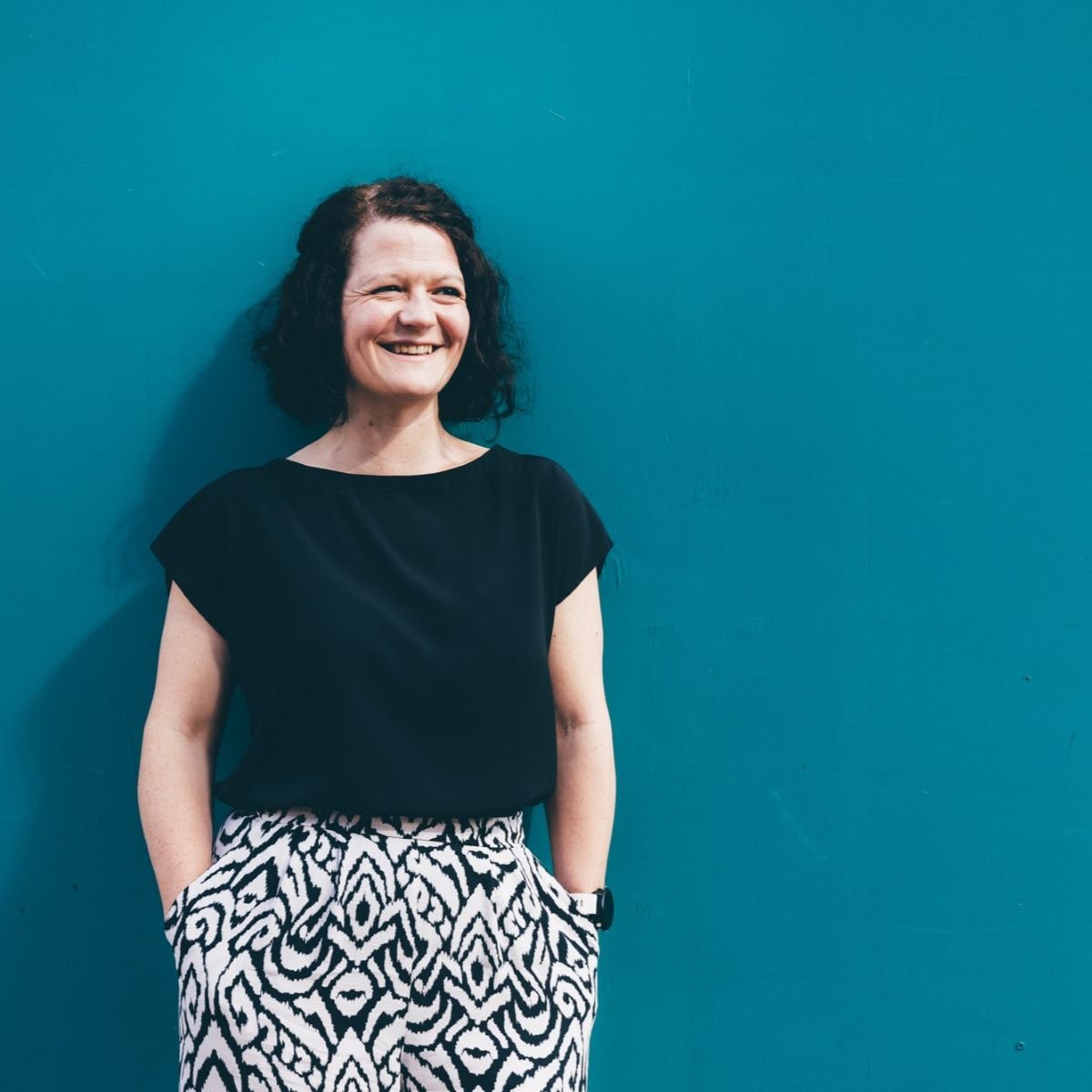 A woman with shoulder length brown hair pictured wearing a black top, standing in front of a bright teal wall. 