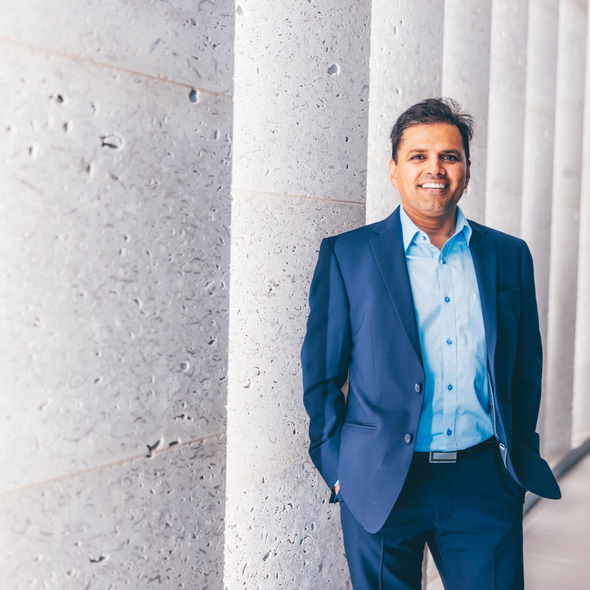 A man is pictured in an office reception, with concrete walls. He is wearing a blue shirt and a dark blue suit jacket, and is smiling at the camera. 