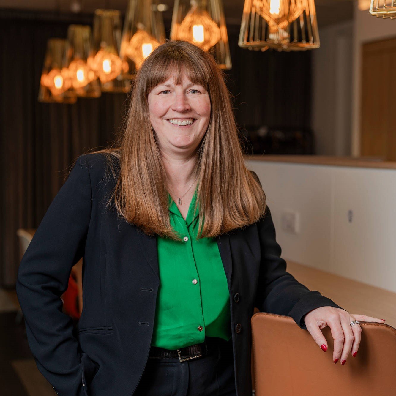 A woman with long brown hair smiles at the camera. She is in an office building and is wearing a green shirt and black blazer.