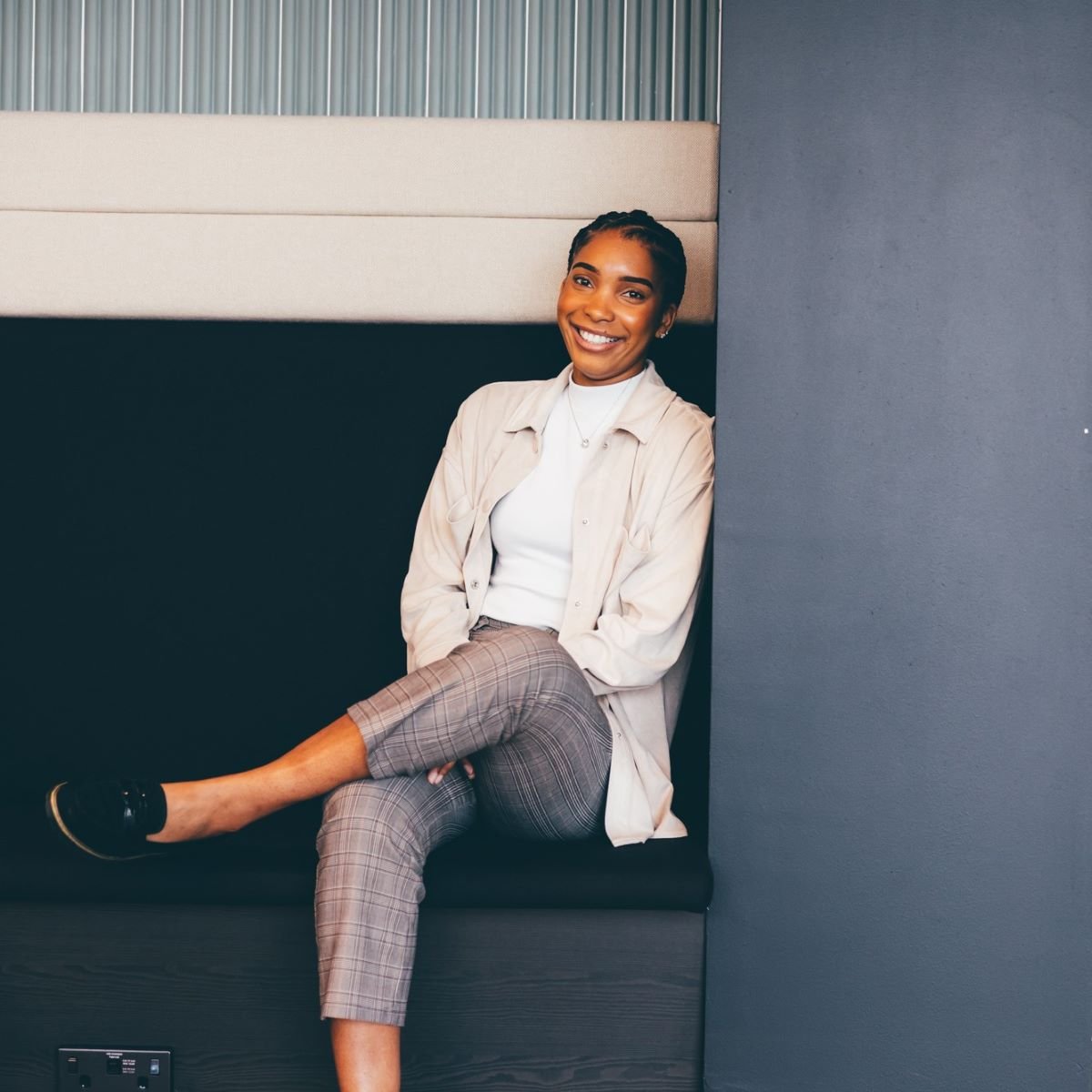 A young woman is pictured sitting on bench seating in an office, she is leaning against a wall. She is looking directly at the camera and smiling, wearing a pale cream top and shirt.