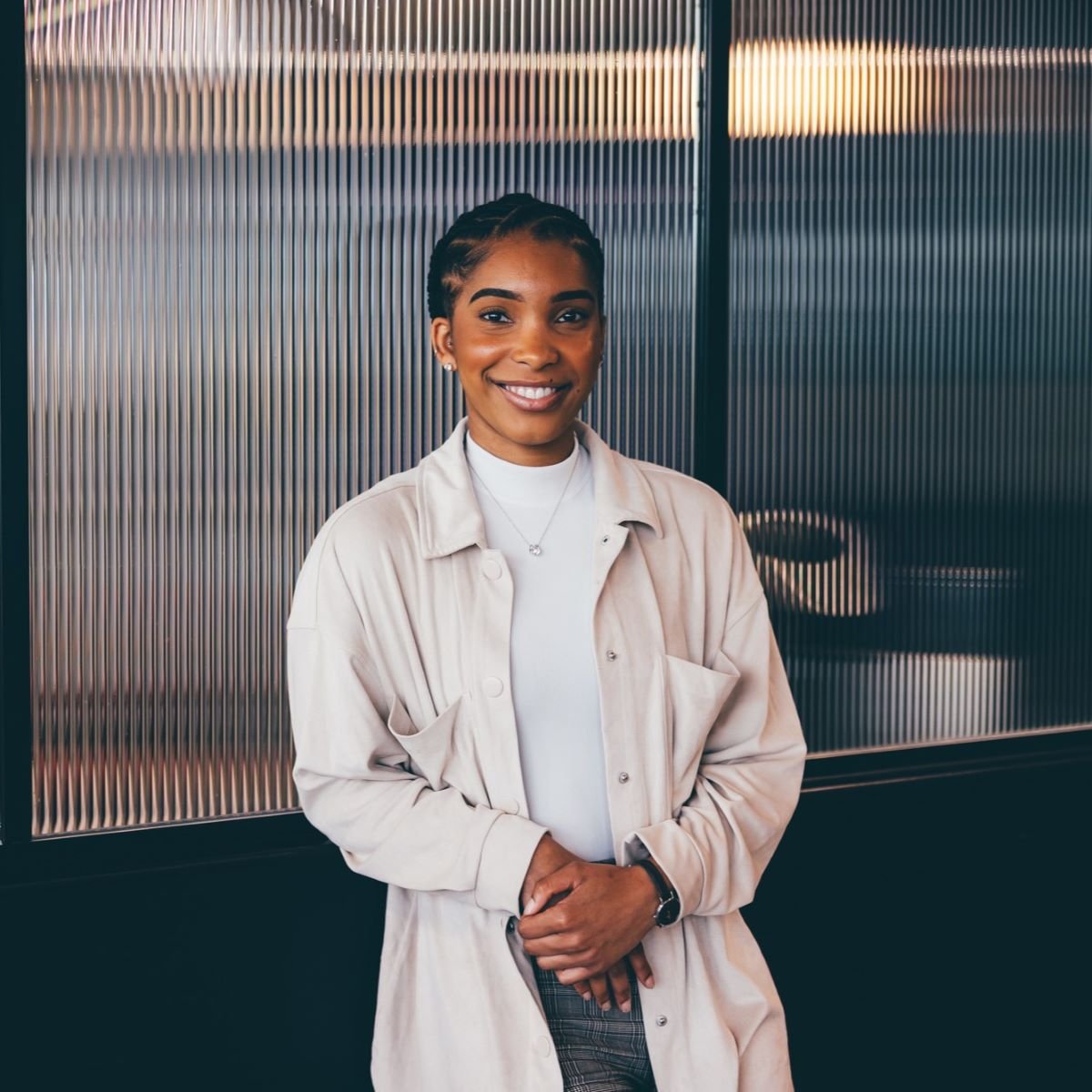 A young woman is pictured in front of a frosted glass panel. She is looking directly at the camera and smiling, wearing a pale cream top and shirt.