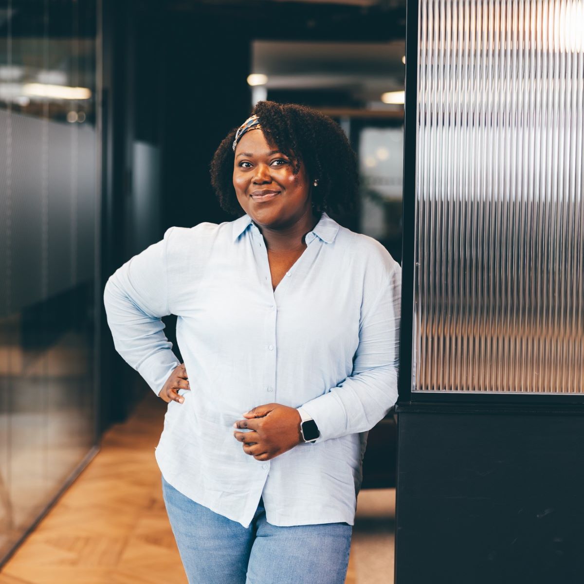 A woman is pictured in an office space, wearing a pale blue shirt and white trousers. She is smiling at the camera with a hand on her hip.