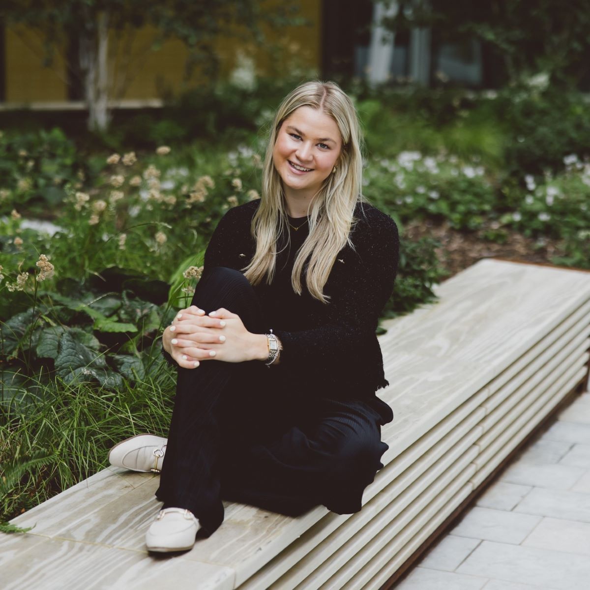 A young woman with long blond hair is photographed wearing a black top, sat on a concrete bench with her hands on her knee. 