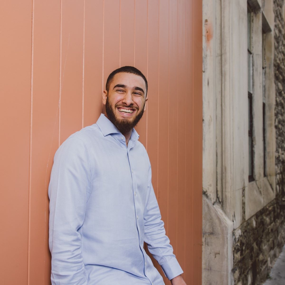 A young man is stood leaning against a peach coloured wooden high wooden door. He is wearing a pale blue shirt, and smiling at the camera, looking to his left.