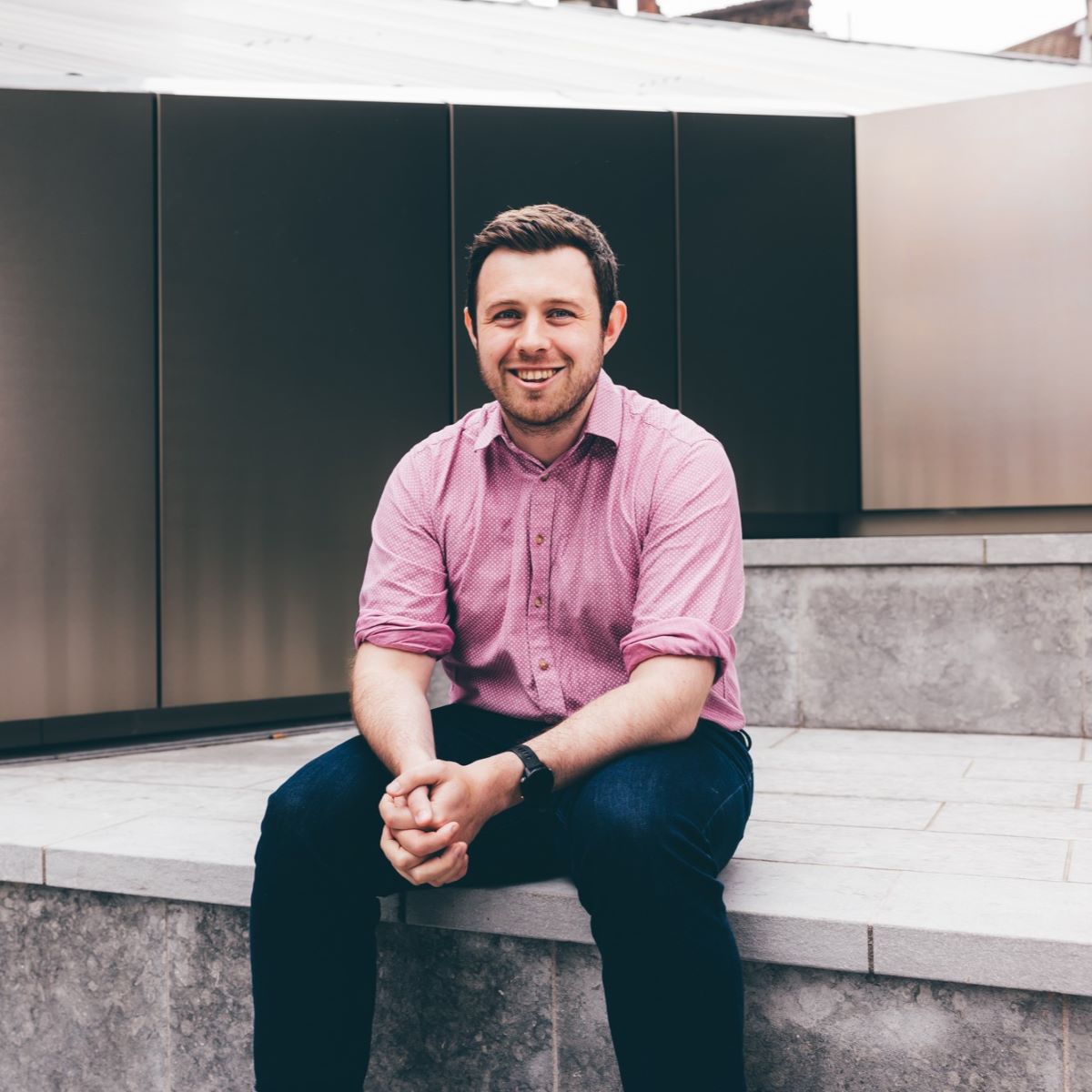 A young man is pictured wearing a pink shirt. He is sat on steps in a courtyard area. 