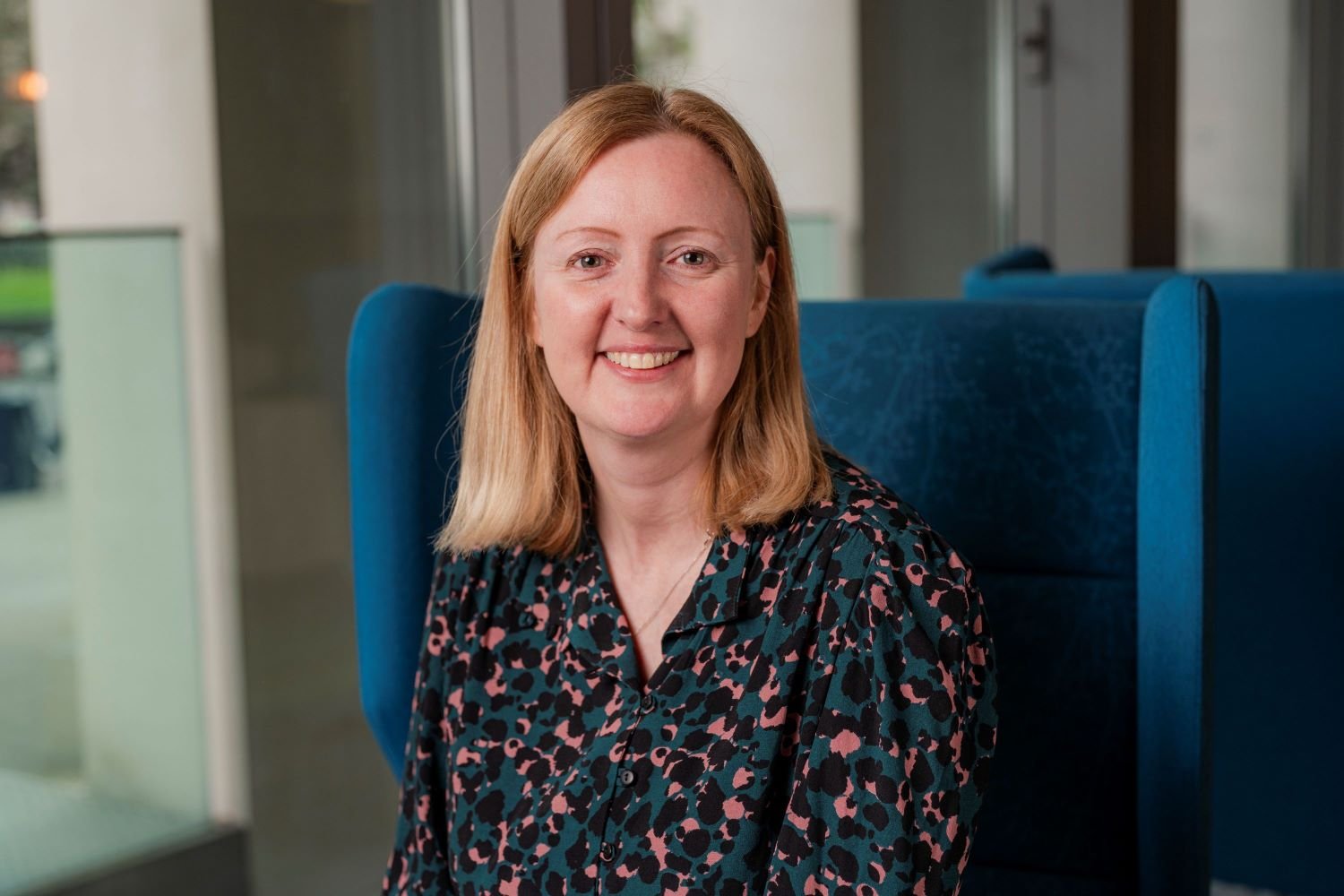 A women with shoulder length blonde hair is sat in a blue chair inside an office, smiling at the camera.