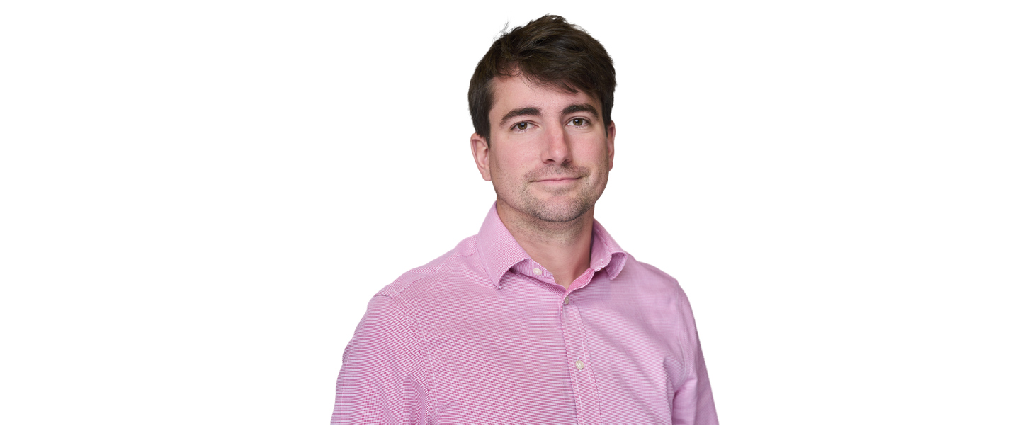 A man with short brown hair stands facing the camera with a smile, wearing a pink collared shirt.