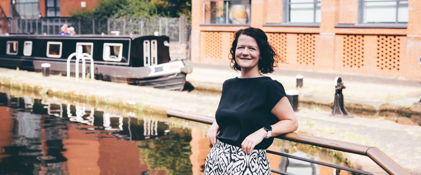 A woman with short brown hair is pictured leaning against railings by a canal. She is wearing a black short-sleeved top and black and white trousers, and is smiling at the camera. 