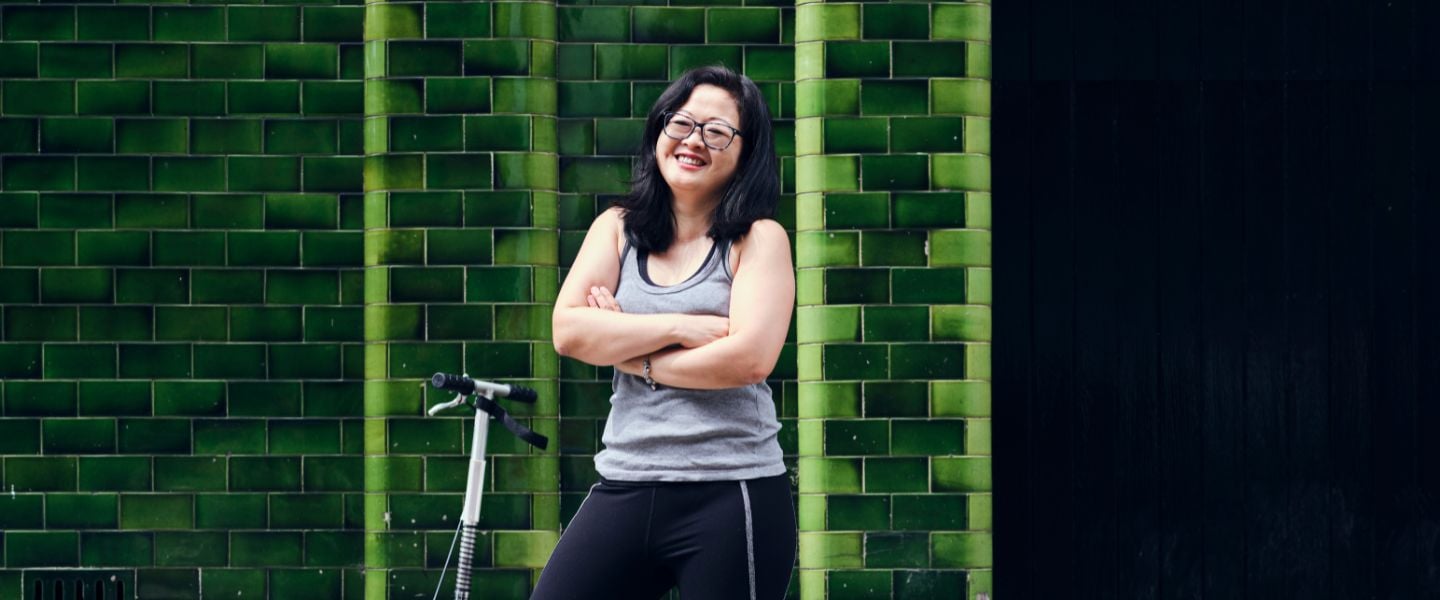 A woman is shown standing in front of a green tiled wall. She is wearing sports wear and is stood next to a scooter. 