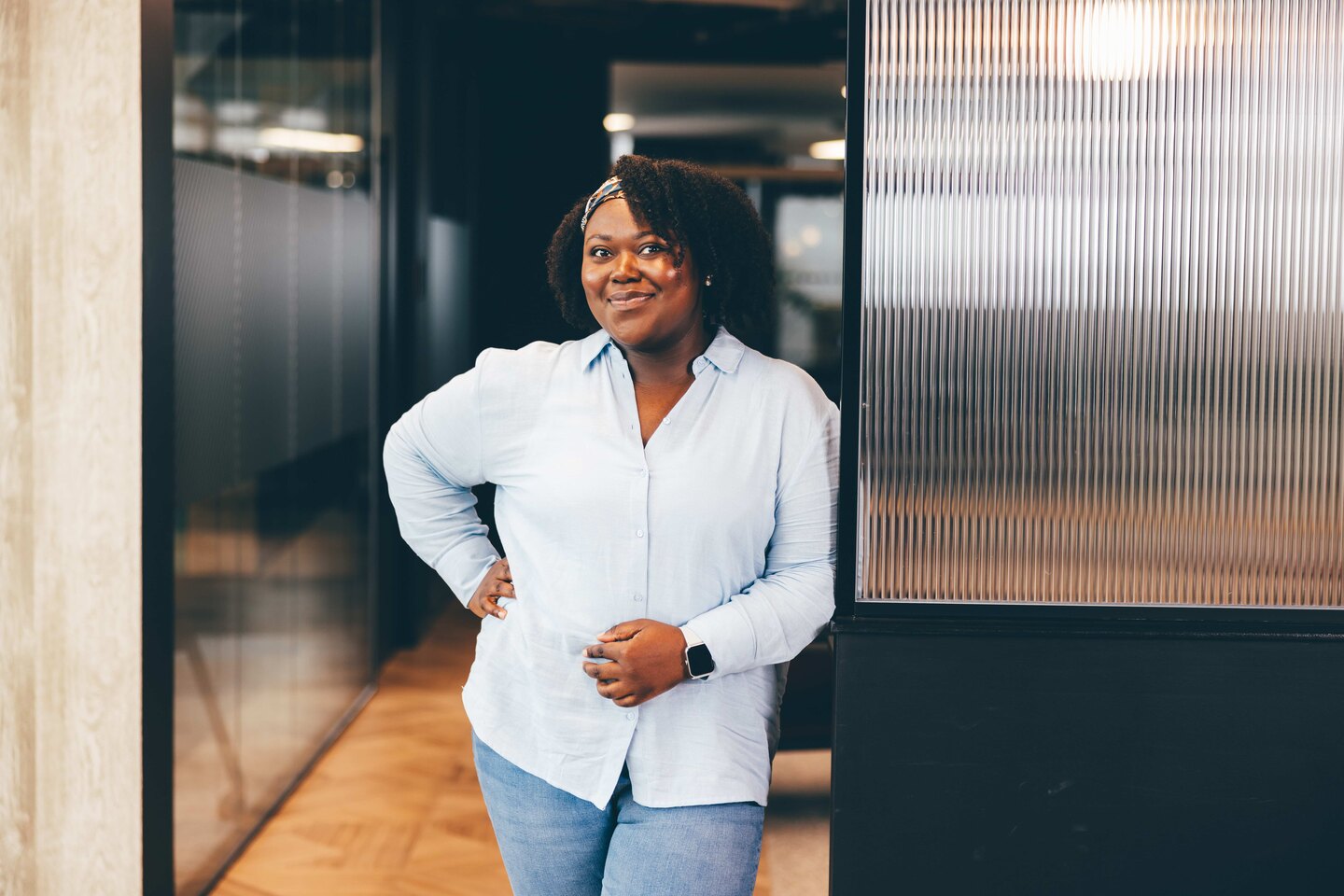 A woman is pictured in an office space, wearing a pale blue shirt and white trousers. She is smiling at the camera with a hand on her hip.