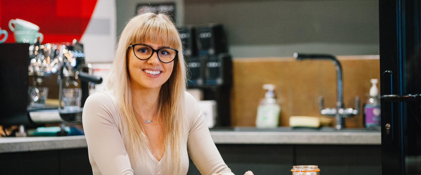 A young woman with blonde hair and glasses is photographed sitting in a coffee shop, smiling at the camera. 