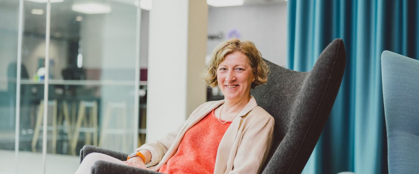 A woman is pictured wearing coral top and beige jacket. She is smiling at the camera, and sitting in a low chair with open office space in the background. 