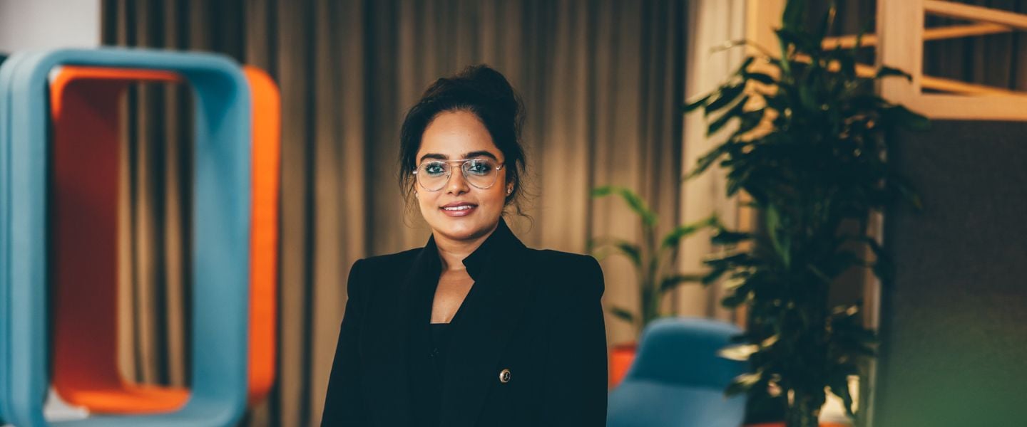 A young woman is pictured wearing a black suit and glasses. She is looking slightly off camera, smiling.
