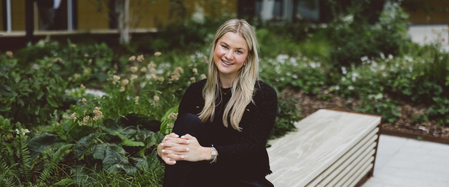 A young woman with long blond hair is photographed wearing a black top, sat on a concrete bench with her hands on her knee. 
