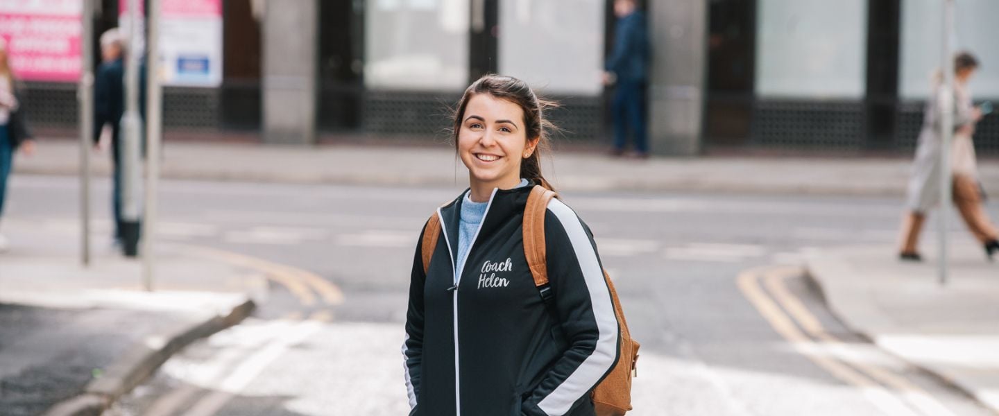 A woman is stood in the middle of a street, wearing a black jacket which says the word coach . She has her brown hair tied back and is smiling at the camera.