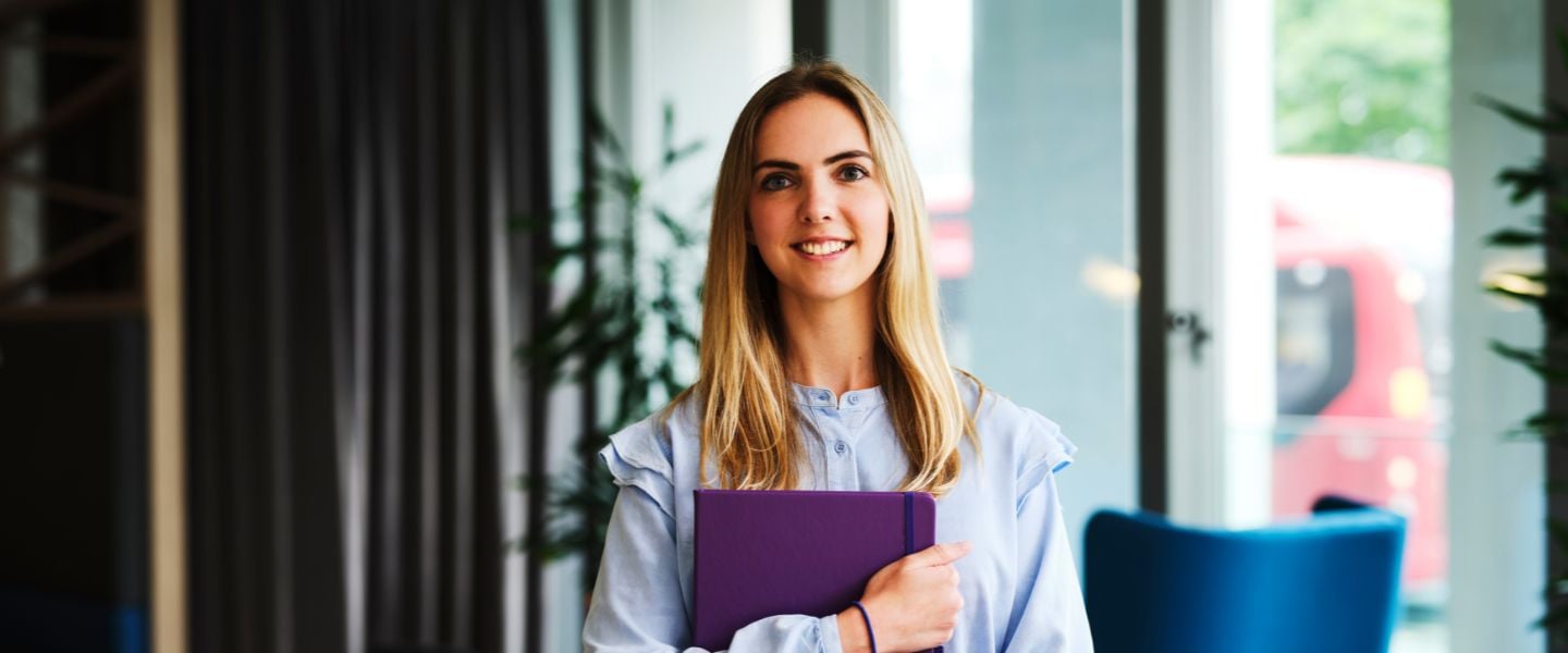 A young woman is photographed wearing an oversized blue shirt. She is standing looking at the camera, and is holding a purple notebook to her chest, in an office environment. 