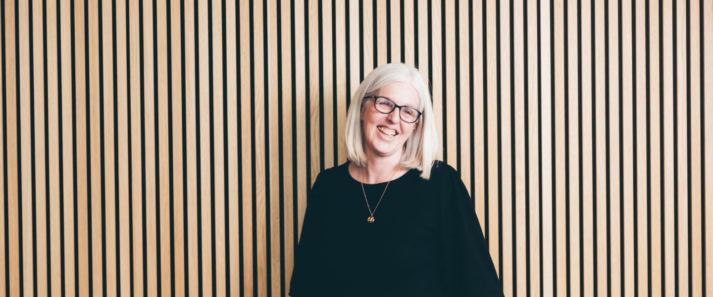 A woman with shoulder length grey hair is standing in front of a wooden slat wall. She is stood facing the camera, laughing, pictured waist upwards wearing a black top. 
