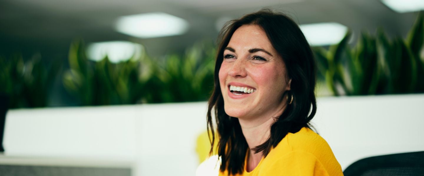 A young woman with brown hair and a bright yellow top is smiling widely whilst looking off camera. She is photographed from her shoulder upwards, from a side angle. 