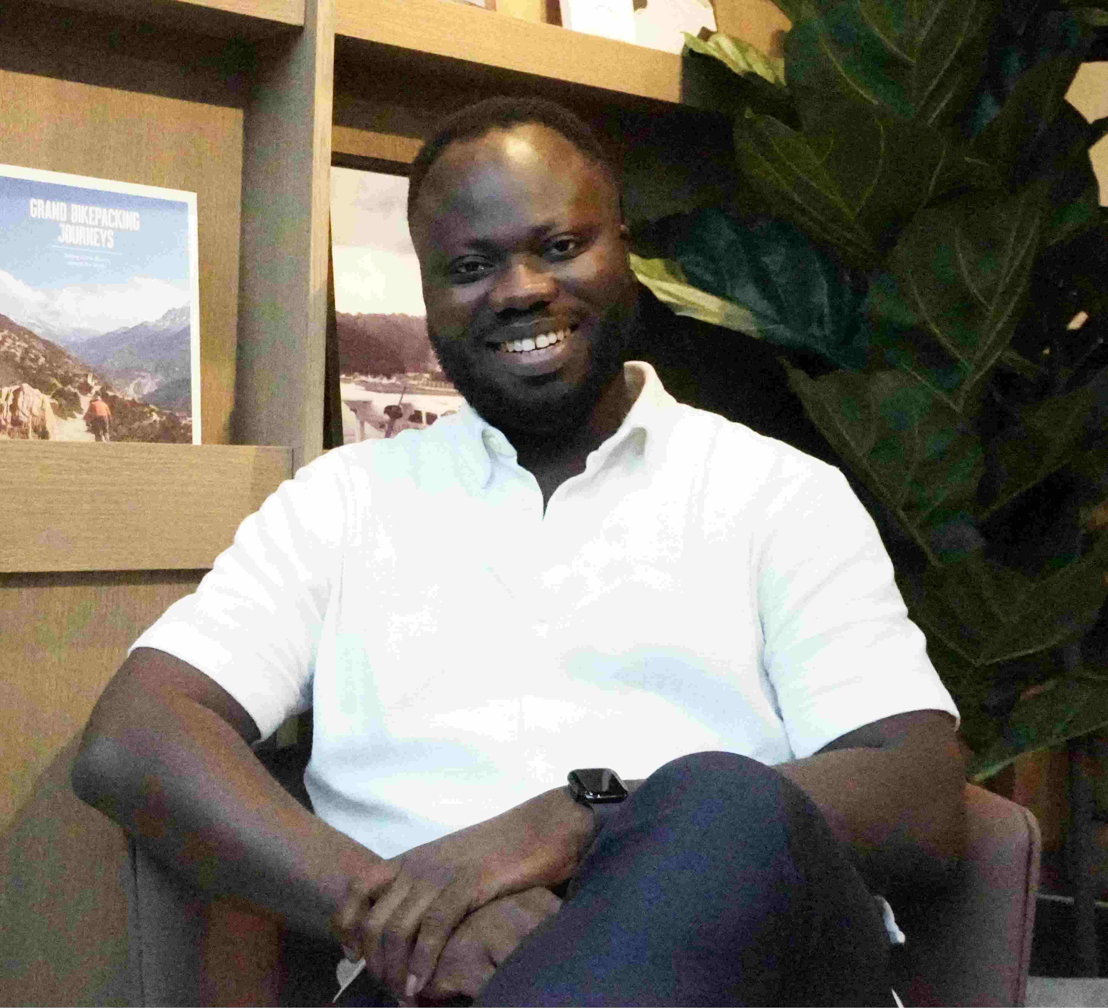 A Black man sits in a chair inside a library, smiling at the camera. He is visible from the waist up. He has short black hair and a beard, and is wearing a white short-sleeved shirt.