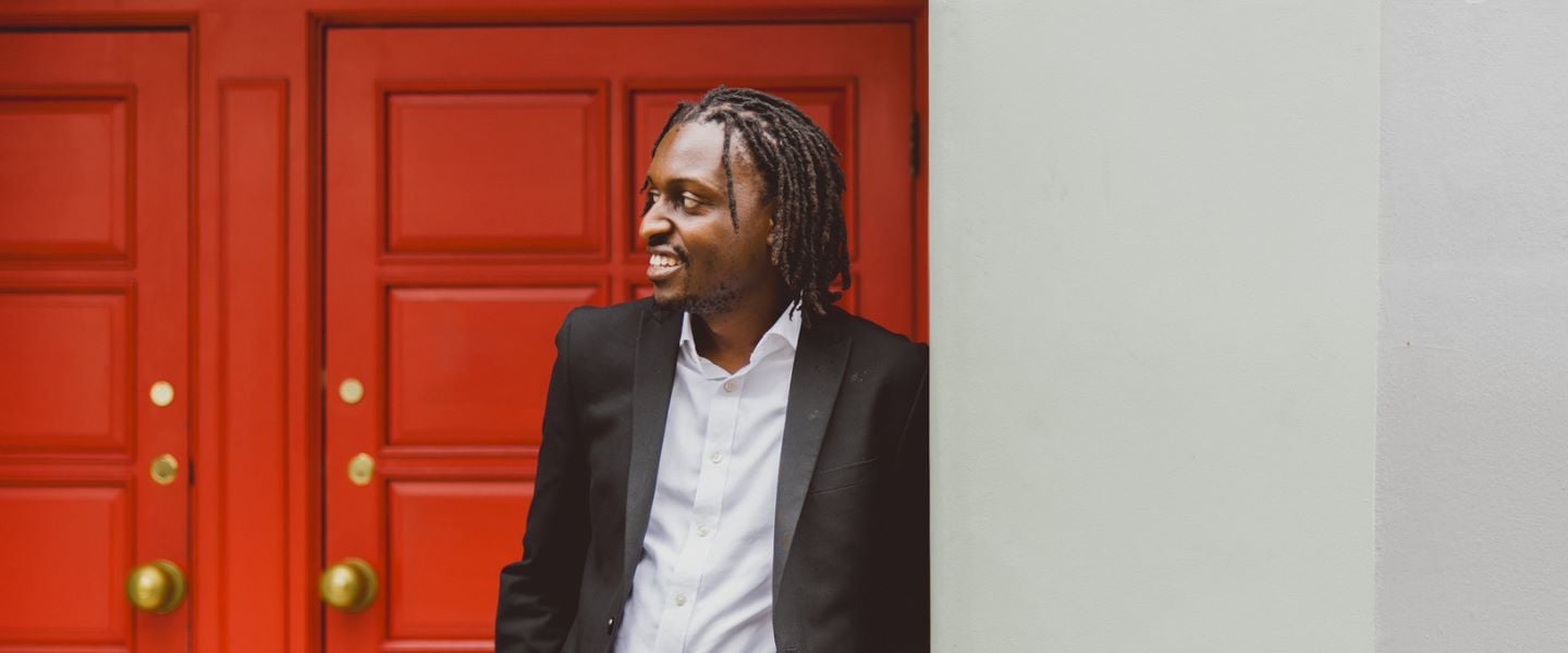 A man is photographed standing in a doorway. The door is bright red, and the wall is pale grey. He is wearing a white shirt and black suit jacket, and smiling whilst looking off camera to the right.