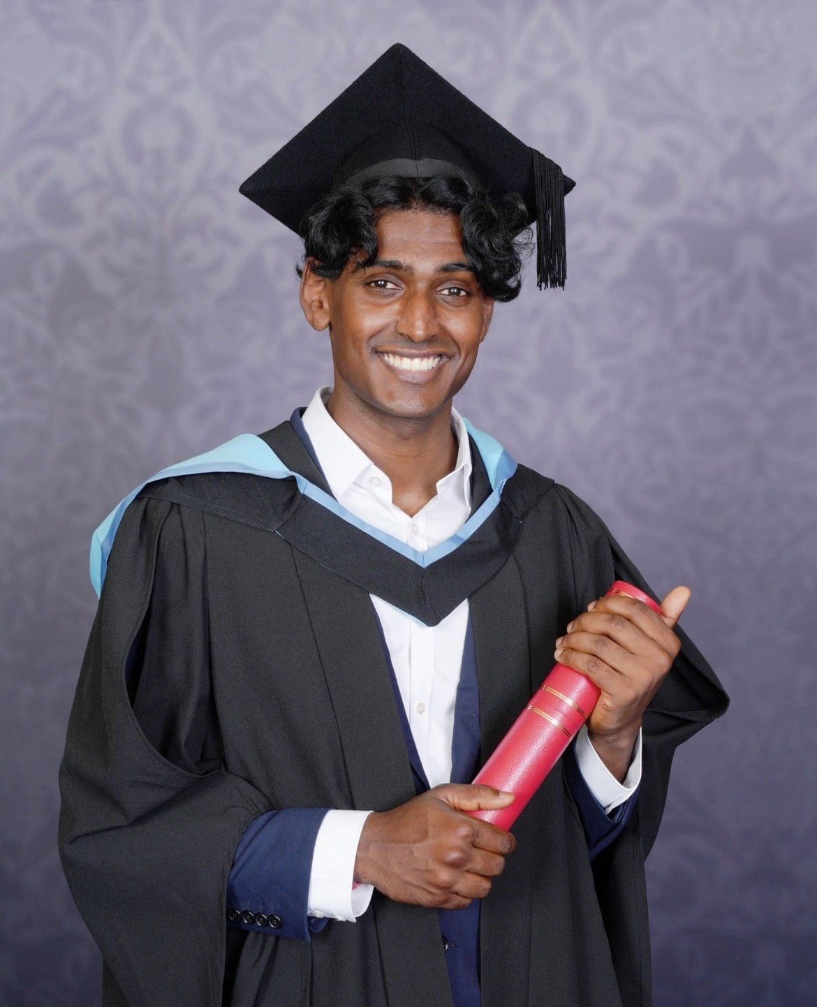 A young South Asian man is posing for a formal and professional graduation photo. He is smiling at the camera, holding his degree, and wearing a cap and robes.