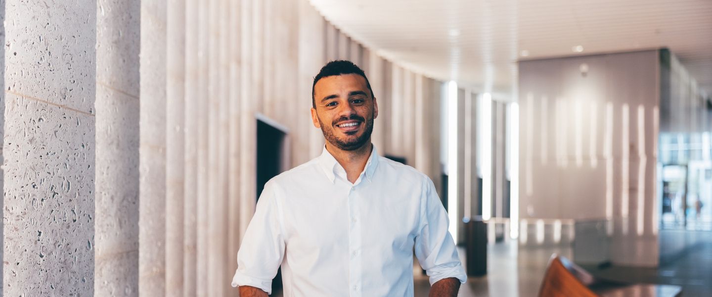 A man wearing a white shirt if pictured in a light and airy office reception area with concrete walls. 