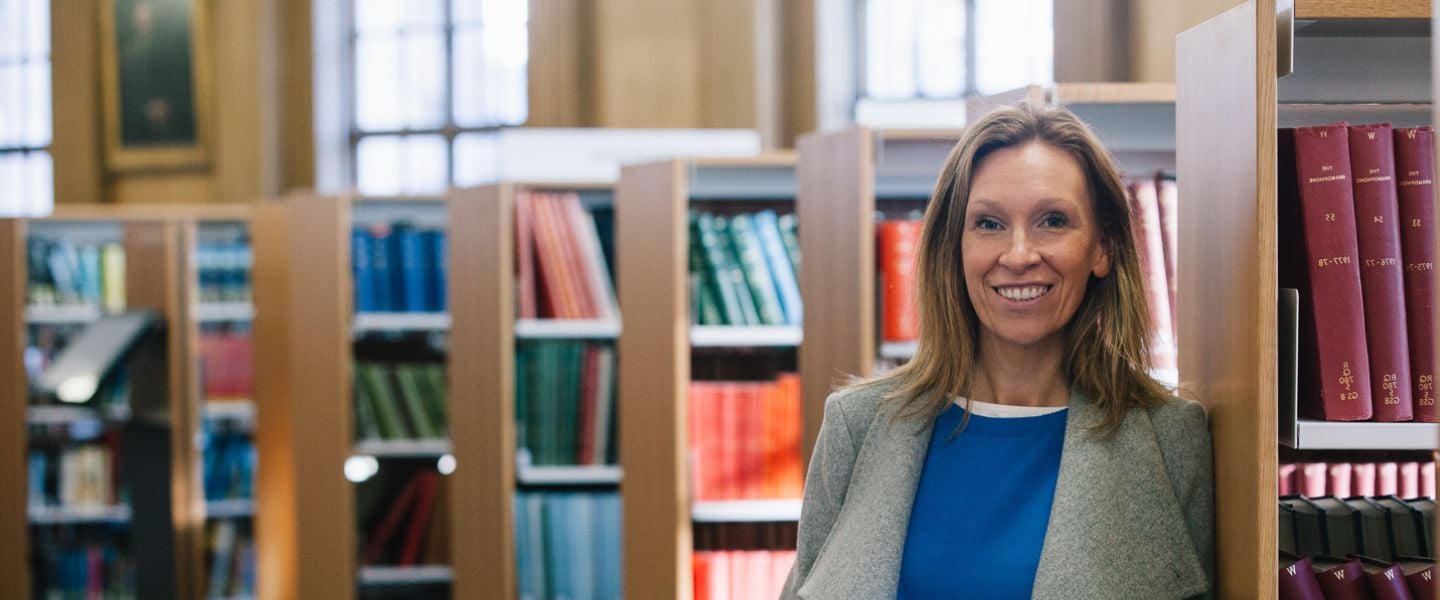 A woman is stood in a library, with rows of books behind her. She is looking straight at the camera, smiling, wearing a grey overcoat and a vibrant blue top. 