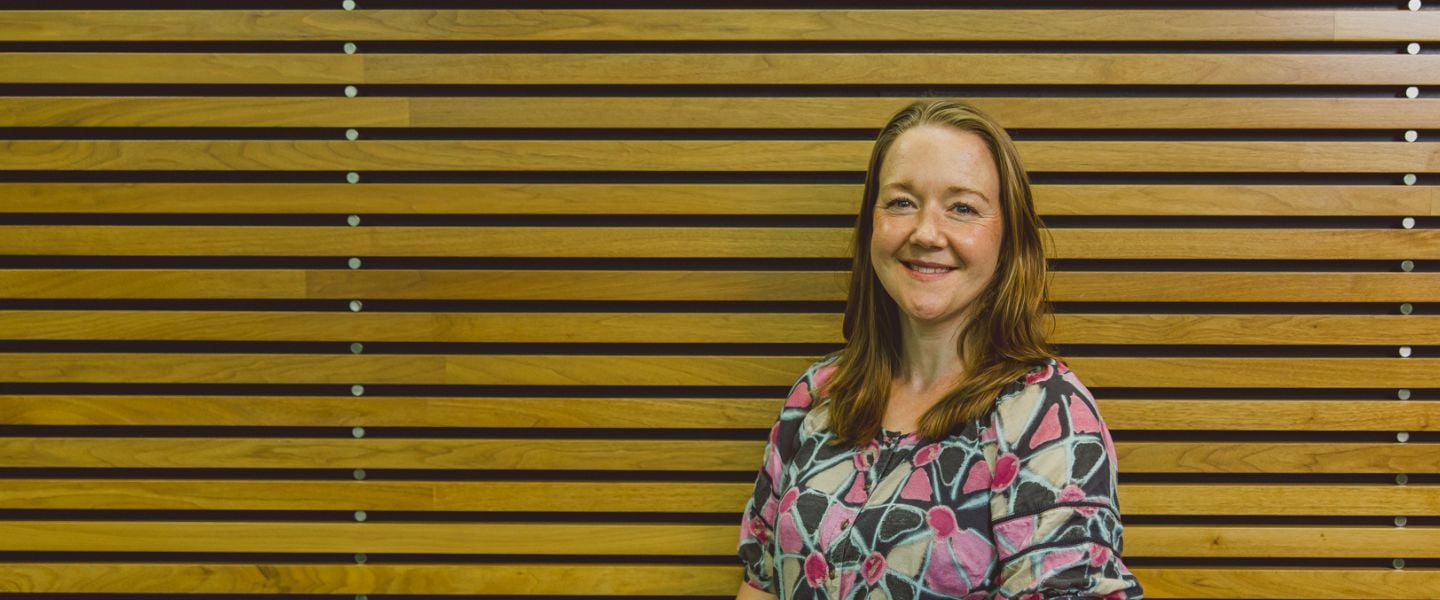 A lady is pictured standing in front of a wooden slat wall. She is smiling at the camera and wearing a grey dress with brightly coloured patterns.