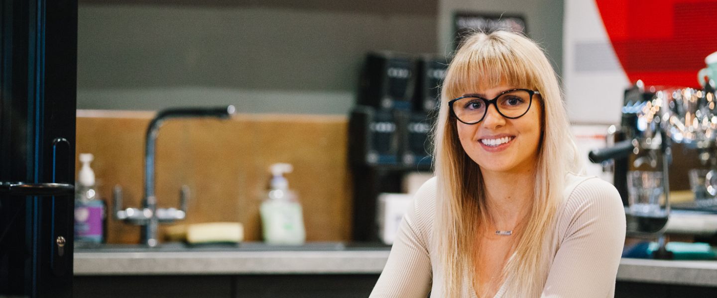 A young woman with blonde hair and glasses is photographed sitting in a coffee shop, smiling at the camera. 