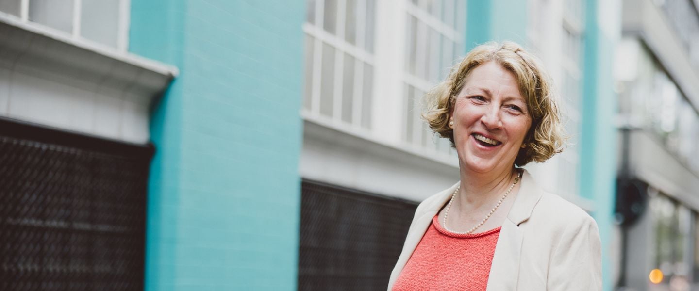 A woman is with short cropped hair, pictured smiling at the camera. She is wearing a coral top and beige jacket, standing in front of a building with a bright teal brick wall and grey shutters.