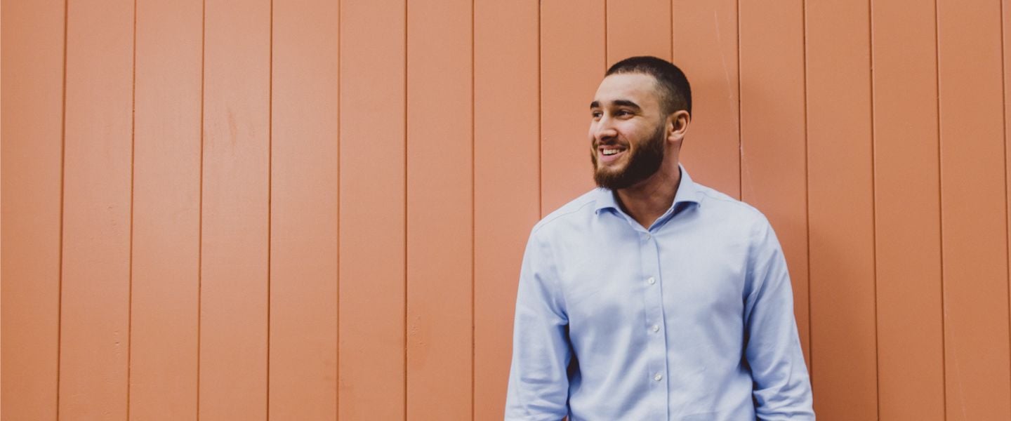 A young man is stood leaning against a peach coloured wooden high wooden door. He is wearing a pale blue shirt, and smiling whilst looking off camera to the right.