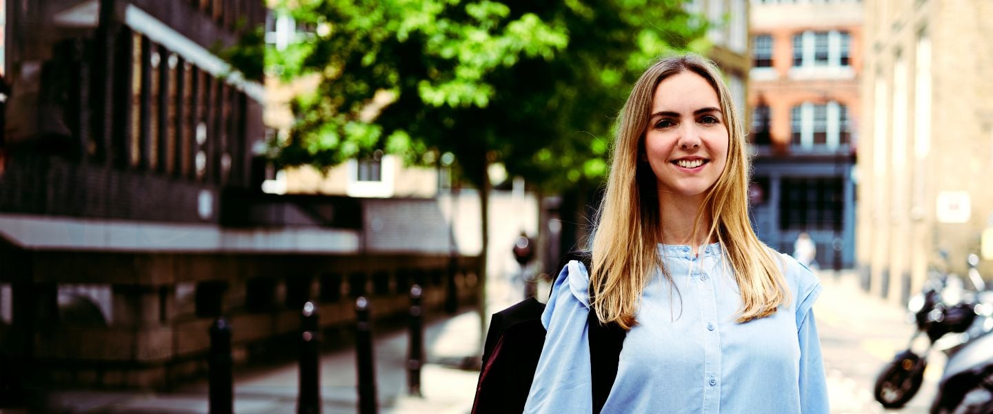 A young woman with blond hair is pictured standing on a street, with a line of mopeds to one side. She is wearing a pale blue shirt, and is carrying a black rucksack on her back. 