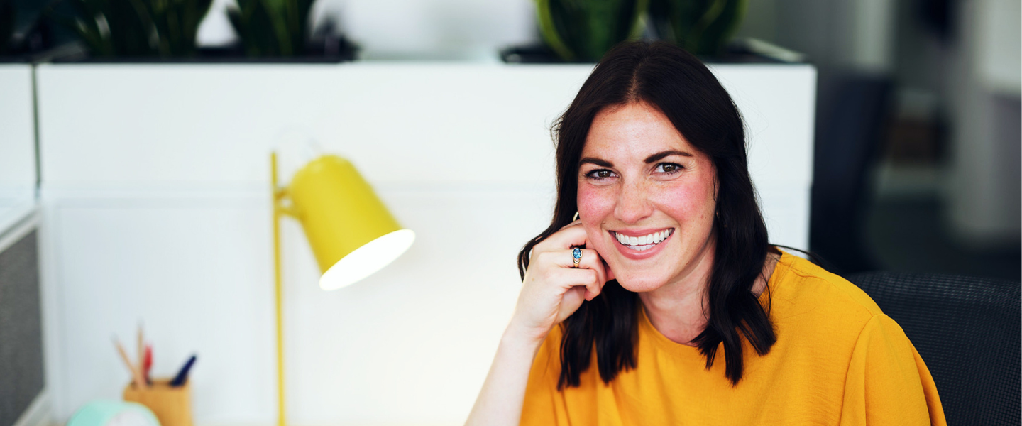 A young woman with brown hair and a bright yellow top sitting at a white office desk, turned towards the camera smiling. 